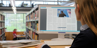 Im Lesesaal der Deutschen Nationalbibliothek liest eine Benutzerin auf dem Computerbildschirm die E-Paper-Ausgabe einer Tageszeitung. Auf dem Tisch liegen einige gedruckte Zeitungsausgaben. 