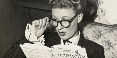 Fotografie einer jungen Frau mit Brille, die überrascht in ein Buch (zu Einsteins Relativitätstherie) schaut