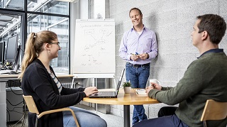 Three employees in a meeting. Two employees are sitting at a table, one employee is standing in front of a flip chart