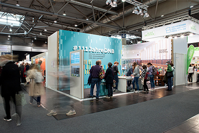 Action area of the German National Library at the Leipzig Book Fair. In the foreground, people passing by can be seen out of focus. In the background the hashtag #111JahreDNB can be read.