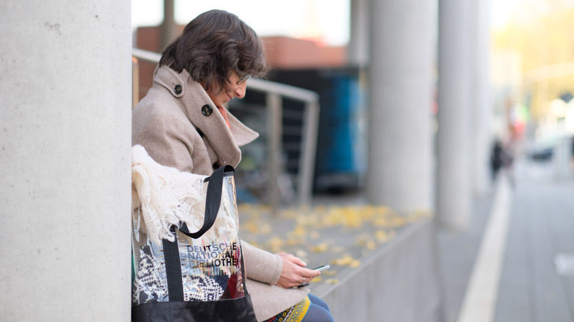 Eine Frau sitzt mit einer Tasche der Deutschen Nationalbibliothek auf einer Betonmauer - und liest in ihrem Smartphone.