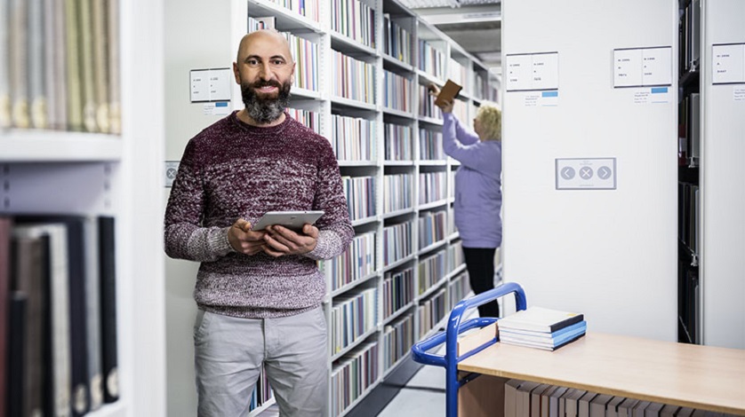 Zwei Beschäftigte im Magazin: Im Hintergrund sortiert eine Mitarbeiterin Bücher in ein Magazinregal. Im Vordergrund steht ein Mitarbeiter mit Tablet in der Hand neben einem Bücherwagen. Zwei Beschäftigte im Magazin: Im Hintergrund sortiert eine Mitarbeiterin Bücher in ein Magazinregal. Im Vordergrund steht ein Mitarbeiter mit Tablet in der Hand neben einem Bücherwagen.