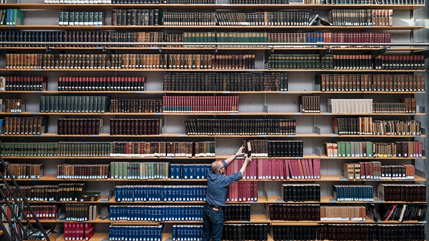 Regalwand voller Bücher, davor steht ein Mann und stellt ein Buch zurück.