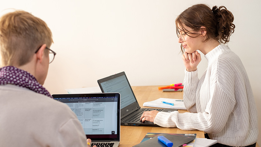 Zwei Frauen sitzen vor ihrem Laptop an einem Schreibtisch mit Arbeitsmaterialien. Zwei Frauen sitzen vor ihrem Laptop an einem Schreibtisch mit Arbeitsmaterialien.