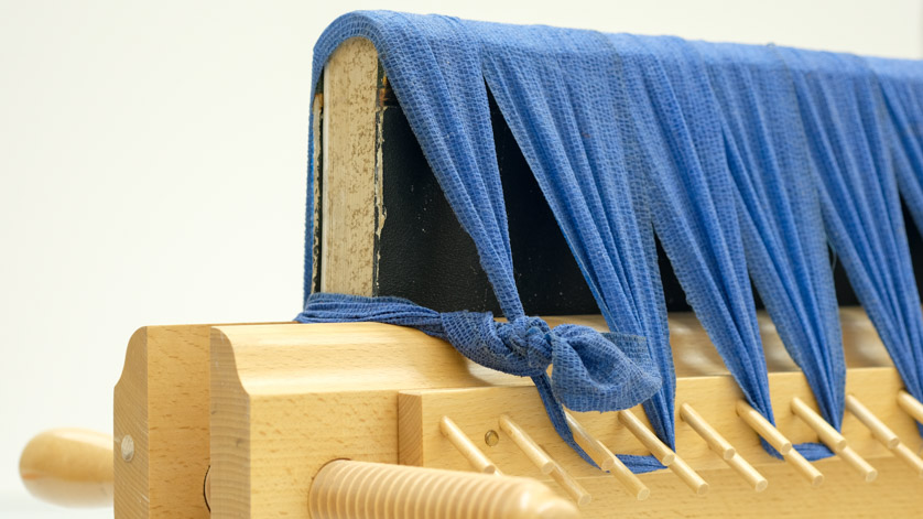 Ein Buch ist in einer Holzpresse fixiert. Dabei wird der angesetzte Rücken des Buches durch eine elastische Binde niedergehalten.