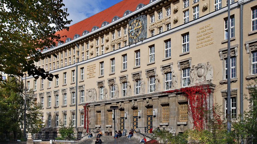 Fassade des historischen Gründungsbaus der Deutschen Nationalbibliothek in Leipzig mit Treppe zum Portal im spätsommerlichen Licht. Menschen sitzen auf der Treppe, rotgefärbtes Weinlaub umrankt die Seiten des Eingangs