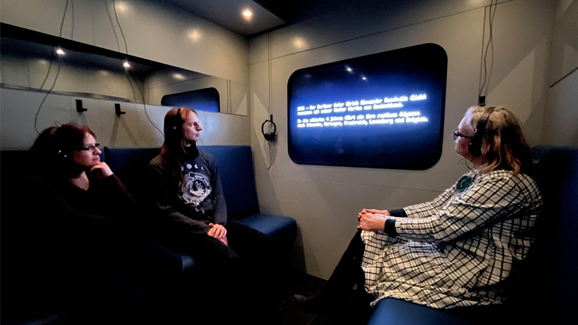 Drei Erwachsene, mit Kopfhöhren auf dem Kopf, schauen auf ein fenstergroßes Display, in einem Raum der einem Zugabteil nachenmpfunden ist. Drei Erwachsene, mit Kopfhöhren auf dem Kopf, schauen auf ein fenstergroßes Display, in einem Raum der einem Zugabteil nachenmpfunden ist.