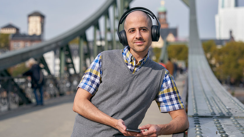  Ein Mann steht auf einer Brücke und hört Musik über Kopfhörer; im Hintergrund ist die Silhouette von Frankfurt am Main zu sehen