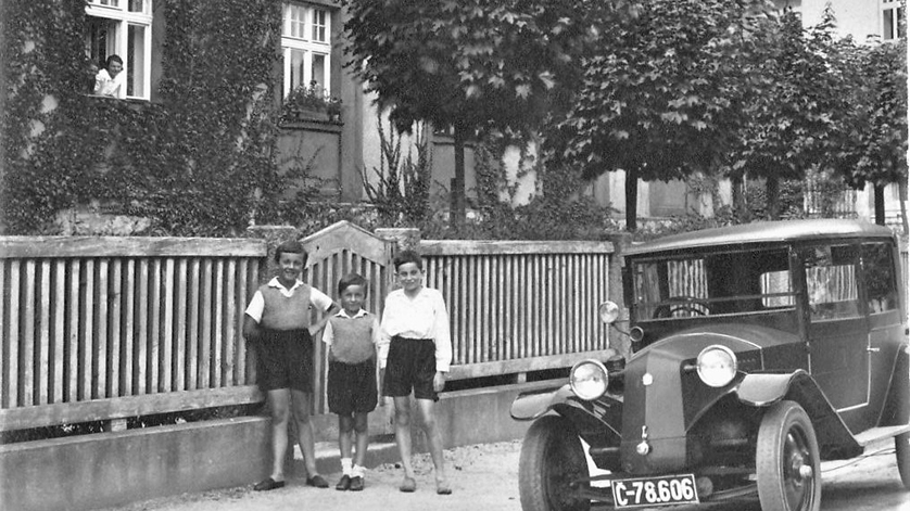 Drei Kinder (Herbert Löwit und die beiden Söhne von Josef Syrowatka) stehen vor einem Gartenzaun, im Vordergrund parkt ein Auto, im Hintergrund schaut eine Frau aus dem Fenster.