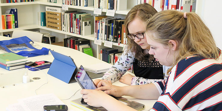Zwei Jugendliche sitzen an einem Tisch mit Büromaterialien und schauen gemeinsam auf ein Laptop. Im Hintergrund ein Bücherregal.