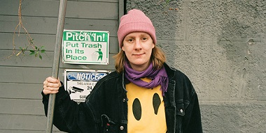 Eine Frau mit pinker Mütze, gelbem Smiley-Pullover und schwarzer Jacke steht vor einer grauen Wand mit zwei Hinweisschildern. Eine Frau mit pinker Mütze, gelbem Smiley-Pullover und schwarzer Jacke steht vor einer grauen Wand mit zwei Hinweisschildern.