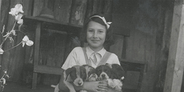 Die Schwarzweißfotografie zeigt Stefanie Zweig im Porträt, auf ihrem Schoß hält sie vier Hundewelpen. 