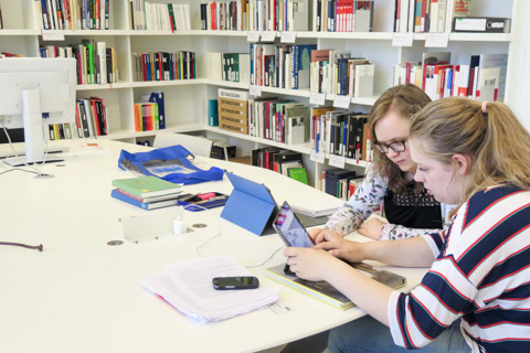 Zwei junge Frauen arbeiten mit Büchern, Ausdrucken und Tablet Computer in einer Bibliothek
