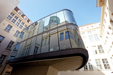 Futuristisch geformter, spiegelnd voll verglaster Gebäudeteil auf einem schmalen Betonsockel in einem historischen Innenhof
