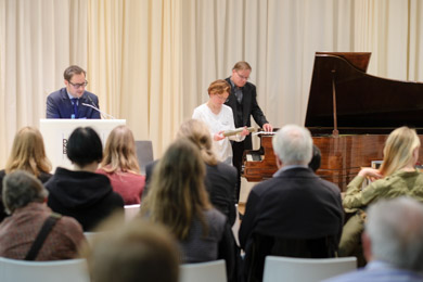 Das Publikum einer Veranstaltung schaut auf das Rednerpult an dem ein Mann vorträgt. Zugleich sind eine Frau und ein Mann an einem Reproduktionsflügel vor dem Publikum damit beschäftigt, eine Klavierrolle auszutauschen Das Publikum einer Veranstaltung schaut auf das Rednerpult an dem ein Mann vorträgt. Zugleich sind eine Frau und ein Mann an einem Reproduktionsflügel vor dem Publikum damit beschäftigt, eine Klavierrolle auszutauschen