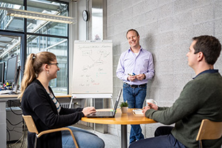 Ein Beschäftigter erklärt zwei Kolleg*innen einen Prozess. Für die Darstellung benutzt er einen Flipchart Ein Beschäftigter erklärt zwei Kolleg*innen einen Prozess. Für die Darstellung benutzt er einen Flipchart