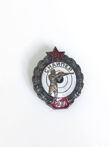 Das runde Metallabzeichen zeigt oben einen roten Stern mit Hammer und Sichel, unten auf rotem Grund den Schriftzug PKKA. In der Mitte ist auf weißem Grund ein bronzefarbener Soldat mit angelegtem Gewehr zu sehen.  