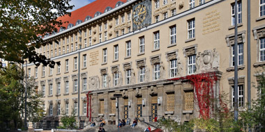 Fassade des historischen Gründungsbaus der Deutschen Nationalbibliothek in Leipzig mit Treppe zum Portal im spätsommerlichen Licht. Menschen sitzen auf der Treppe , rotgefärbtes Weinlaub umrankt die Seiten des Eingangs