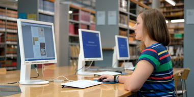 A user reading an online publication at one of our reading room PCs. In the background library shelves with books. A user reading an online publication at one of our reading room PCs. In the background library shelves with books.
