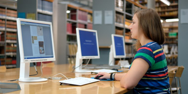 A user reading an online publication at one of our reading room PCs. In the background library shelves with books.