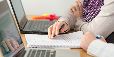 Two people are each sitting in front of a laptop and are reading through notes on a notepad together. Two people are each sitting in front of a laptop and are reading through notes on a notepad together.