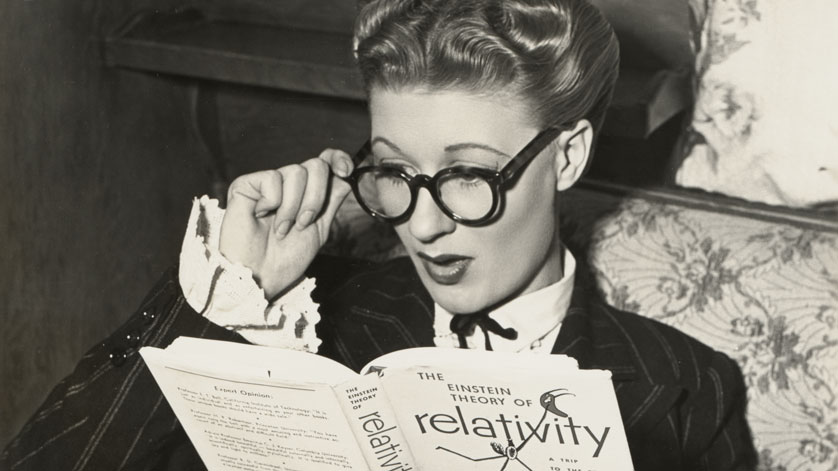 Photo of a young woman wearing glasses and gazing at a book (on Einstein’s theory of relativity) with a look of surprise on her face