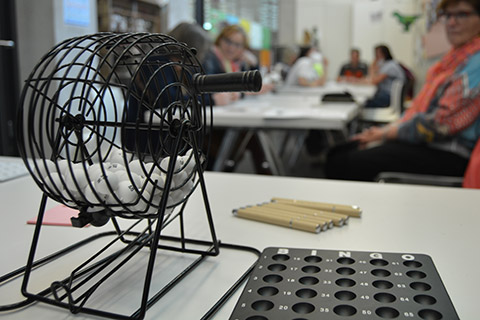 People are sitting at a table playing museum bingo. In the foreground, there is a bingo drum and a number board.  People are sitting at a table playing museum bingo. In the foreground, there is a bingo drum and a number board.