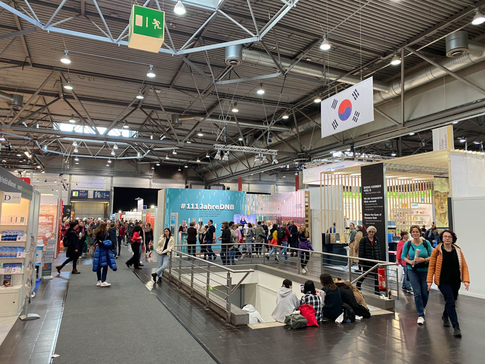 Hustle and bustle at the Leipzig Book Fair 2023 with the German National Library's promotional area in the background and a staircase going down to the basement in the foreground.