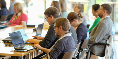 Frauen und Männer sitzen mit Notebooks an Tischen in einem Konferenzsaal und hören einem Vortrag zu Frauen und Männer sitzen mit Notebooks an Tischen in einem Konferenzsaal und hören einem Vortrag zu