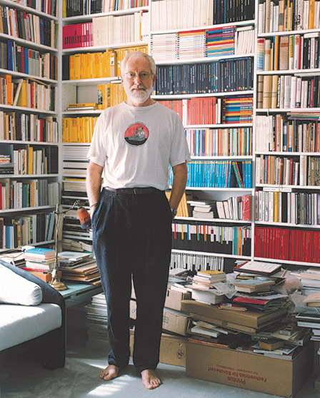 A man standing barefoot in a living room with numerous books in the background and on the floor A man standing barefoot in a living room with numerous books in the background and on the floor