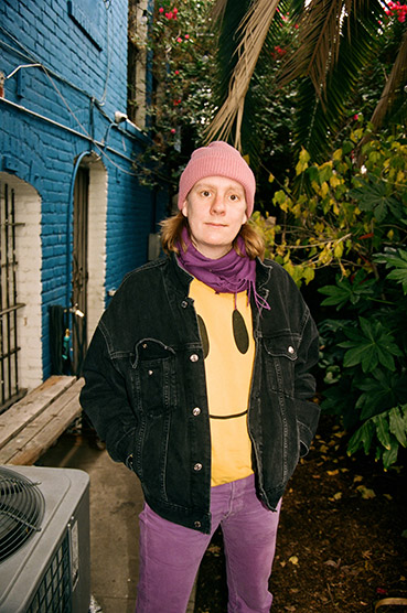 A woman wearing a pink hat, a yellow smiley pullover, purple trousers and a black jacket standing in front of some trees and a brick building painted blue and white. A woman wearing a pink hat, a yellow smiley pullover, purple trousers and a black jacket standing in front of some trees and a brick building painted blue and white.