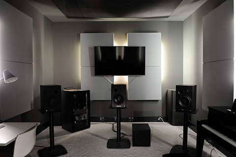 IView inside a room with soundproofing elements on the walls and a large screen. There is a narrow workstation with a monitor on the left side. On the right, there is a digital grand piano.  