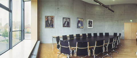 Ovaler, schwarzer Tisch mit 16 Stühlen, seitlich große Fensterfront, an der gegenüberliegenden Wand Portraits früherer Generaldirektoren der Deutschen Nationalbibliothek in Frankfurt  