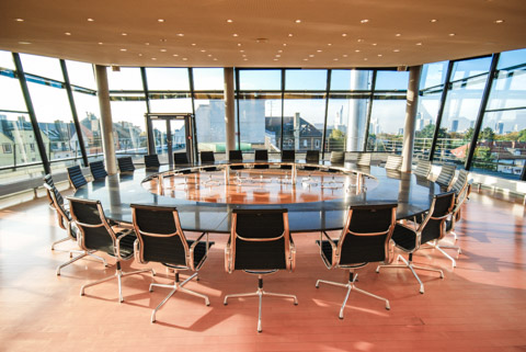 Kreisförmiger Tisch mit Steintischplatte inmitten eines Dachgeschossraumes mit drei vollverglasten Seiten. Um den Tisch 24 Stühle, im Hintergrund die Skyline Frankfurts und eine Dachterrasse 